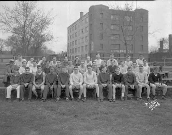 Group portrait of Central High School track team taken at Breese Stevens field, with the Simon Brothers wholesale grocers building (901 E. Washington Avenue) in the background.