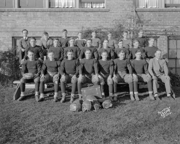 Outdoor group portrait of the Middleton High School football team and coach, sitting on bleachers. Football helmets are in a pile in front of the group.
