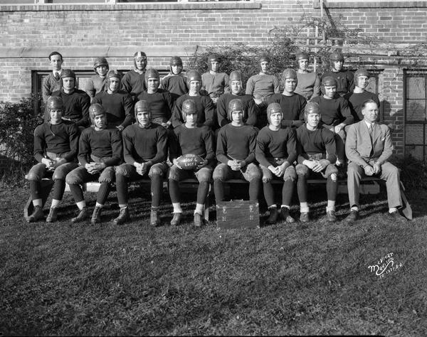 Outdoor group portrait of Middleton High School football team and coach, sitting on bleachers. The team is wearing helmets.
