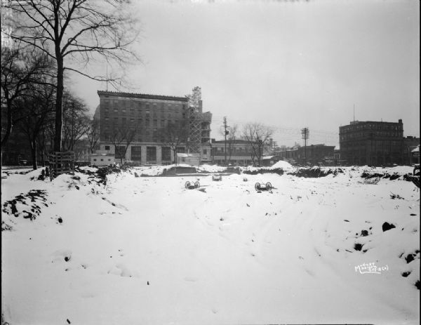 Ground being prepared for the construction of the U.S. Post Office, 215 Monona Avenue, showing Beaver Building (National Mutual Benefit Insurance), 119 Monona Avenue; Cantwell Building, 121 S. Pinckney; Wisconsin Auto Licensing, 16 E. Doty Street and other buildings along E. Doty Street and S. Pinckney Street. Snow is on the ground.