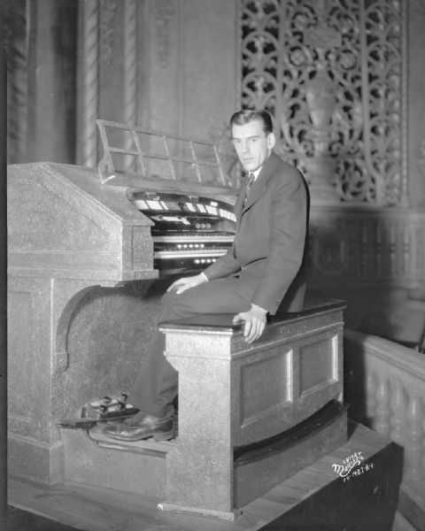 A male organist is sitting on a bench and looking towards the camera in front of the Eastwood Theatre organ. The organ grill is in the background on the far wall. The theatre is at 2090 Atwood Avenue.
