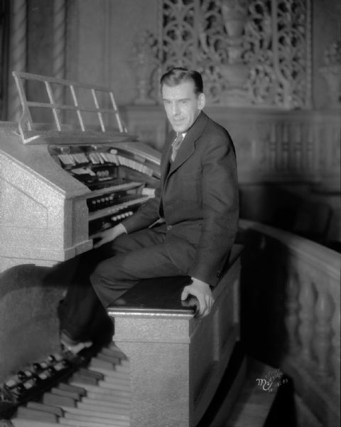 A male organist is sitting on a bench and looking towards the camera in front of the Eastwood Theatre organ. The organ grill is in the background on the far wall. The theatre is at 2090 Atwood Avenue.