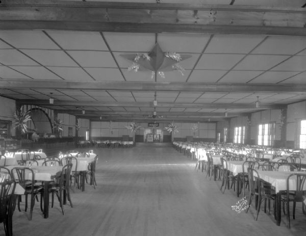Interior view of ballroom of Chanticleer Road House. Tables and chairs are set around the large rectangular room. A row of windows is along the right wall. There is an arched stage on the left in the background.
