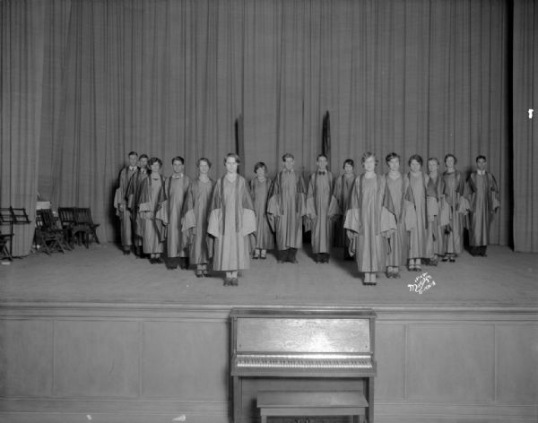 East High School Honor Society students standing on a stage in keystone formation. They are wearing robes. There is a piano in front of the stage.