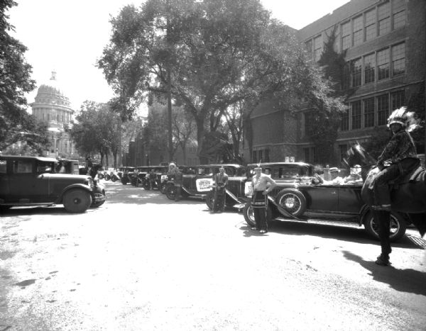 A man on a horse with Pontiac cars lined up on Wisconsin Avenue for the US Indian Band parade with State Capitol in the background. They played a concert at the Orpheum Theatre.