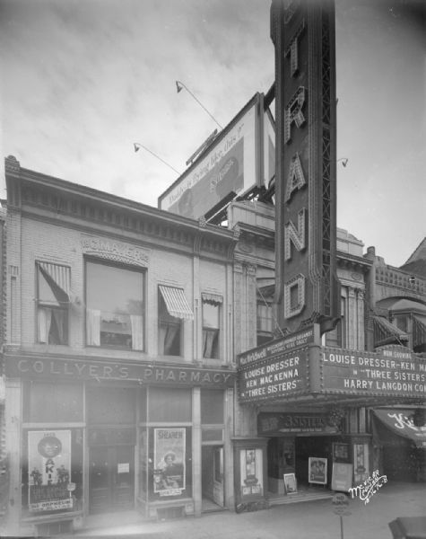 Orpheum advertisement for "The Sap from Syracuse," and a Capitol Theatre advertisement for "Let Us Be Gay" in Collyer's Pharmacy window in the Mayer Building at 14 E. Mifflin Street next to the Strand Theatre. The Strand marquee has a sign for "Three Sisters," at 16 E. Mifflin Street.