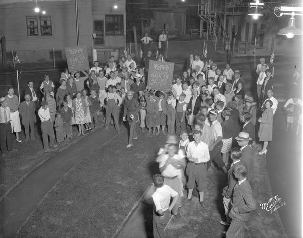 Elevated view of a man in uniform leading a small parade of children who are holding a sign advertising the movie: "Don't miss the year's greatest laugh hit! Joe Cook in Rain or Shine, now playing RKO Capitol" with a small crowd watching.