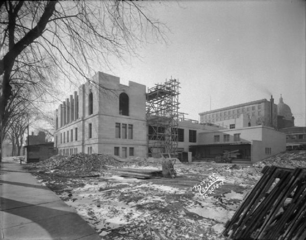 Stone walls are going up on the second floor. The view is looking west at the back of the building, with a wheelbarrow and scaffolding in the lot. In the background is the Beavers Insurance Building, 119 Monona Avenue and the Wisconsin State Capitol.