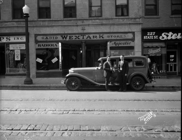 View across street towards two men standing at the side of the Capital Times car in front of the Wextark store, 225 State Street. Stewart Smart Shop is on the right.
