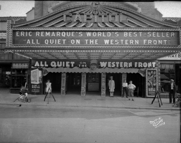 Capitol Theatre marquee at 209 State Street reads: "Eric Remarque's World's Best-Seller 'All Quiet on the Western Front,'" with usher and several bystanders in front of the entrance. Firearms are stacked in tripod formations on the sidewalk, flanking the entrance on the left and right. Two men, one on the left and one on the right, are standing or crouching near devices on tripods, perhaps machine guns.