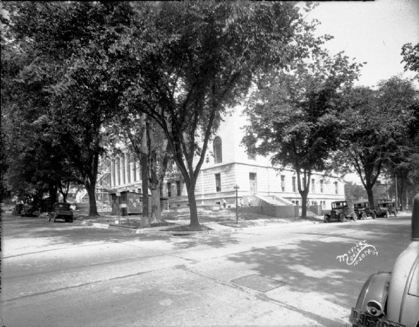 View looking north from corner of Monona Avenue and Wilson Street of front and side of the building, with workmen finishing the basement level. Automobiles are parked along the curbs. There is a construction shack on the left.