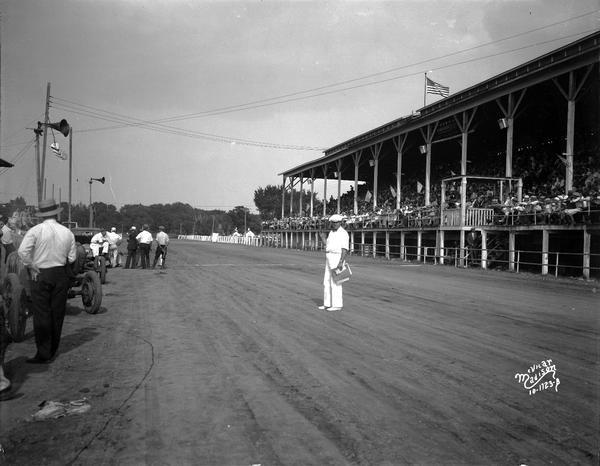 People in grandstand are overlooking the track from the right. There is a man standing in the center of the track making announcement over a public address system. Other men are standing along the left.