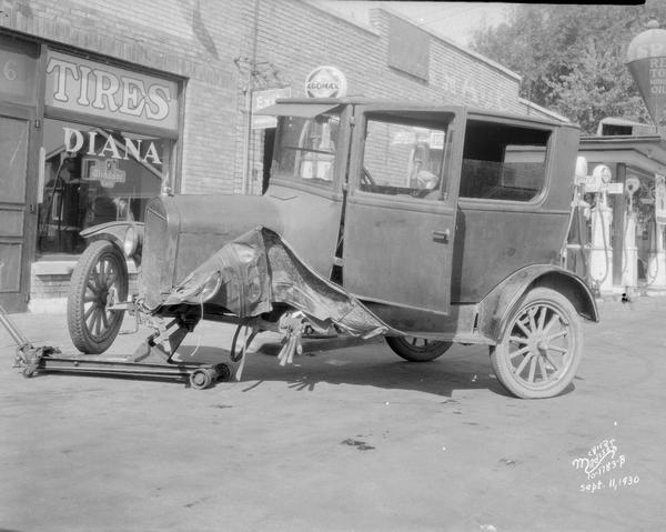 A damaged Ford Sedan automobile at South Park Street garage, 802-806 S. Park Street, with "Tires-Diana" on the storefront window in the background.