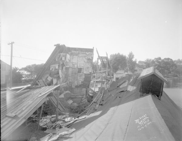 Collapsed ice house in Pardeeville. A roof and debris are in the foreground, and trees and buildings are in the background.
