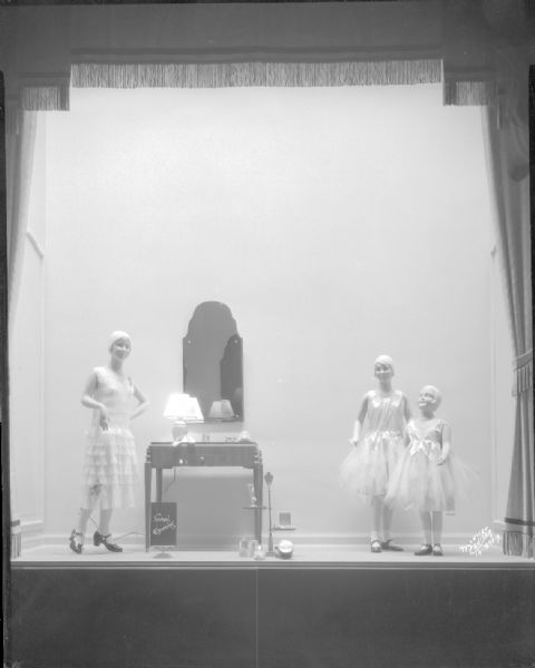 Manchester display window showing three girl mannequins wearing dresses standing near a dressing table and mirror.