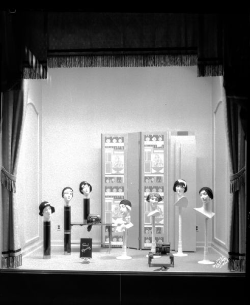 View of the Manchester display window featuring seven mannequin heads with women's hats.