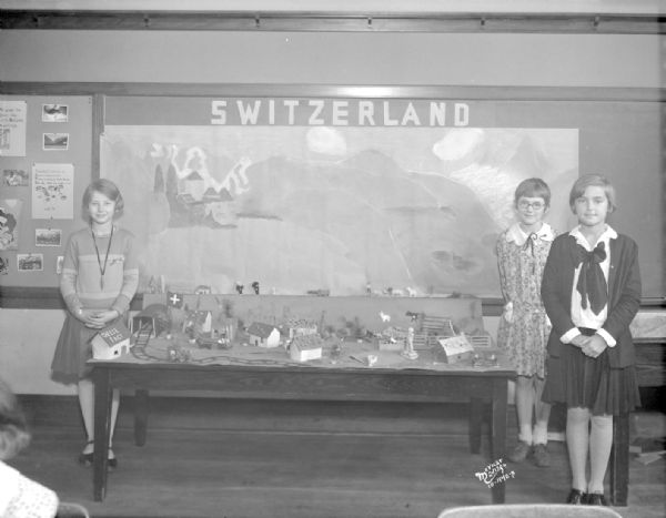 Three girls are standing beside a model they constructed of Switzerland on a table at Randall School.