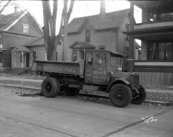 View across street towards a J.B. Drives Fuel Company "old" dump truck made by Diamond T parked along a curb in front of houses.