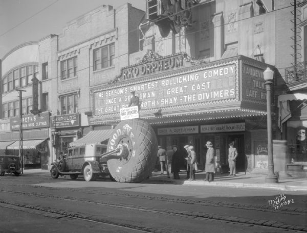 World's largest Goodyear tire in front of RKO Orpheum theater, 216 State Street, with six people viewing the tire. One man is on top of the tire holding a sign. Theater marquee is advertising "Once a Gentleman."  Also shows two stores, Harrold's Credit Jewelers and Speth's Clothing Store.