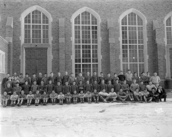 Group portrait of East High School football squad in uniform, taken in front of gymnasium windows.