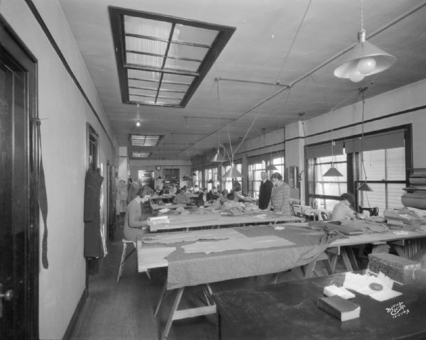 View of the interior of Tiffany's dress making shop (Creators of Claire Tiffany Frocks, 546 State Street), showing women cutting and sewing dresses at long work tables. There is a desk in the foreground, and a man is standing along the windows on the right side of the room.