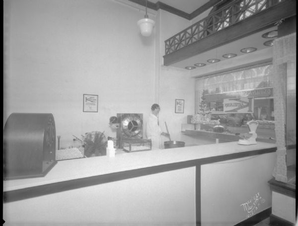 View of the interior of the O'Sullivan's Karmelkorn Shop, 308 North Henry Street, with a small Christmas tree with tinsel on display in the front window on the right. Behind the counter a woman is using a large utensil to stir something in a large pot. A radio is on the counter on the far left.