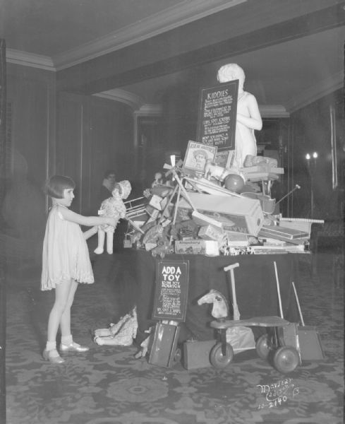 View of a Christmas Add-a-Toy Mountain in the RKO Orpheum theater lobby, with a girl adding a doll to the mountain of toys. The toys are on and around a table in front of a statue of a boy with a goat on a pedestal. Sponsored by the Wisconsin State Journal Empty Stocking Club.