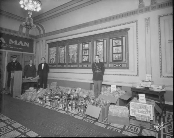 Collection of food for Salvation Army. Shows four men, two in uniform, and two wearing suits, in the Strand theater lobby.
