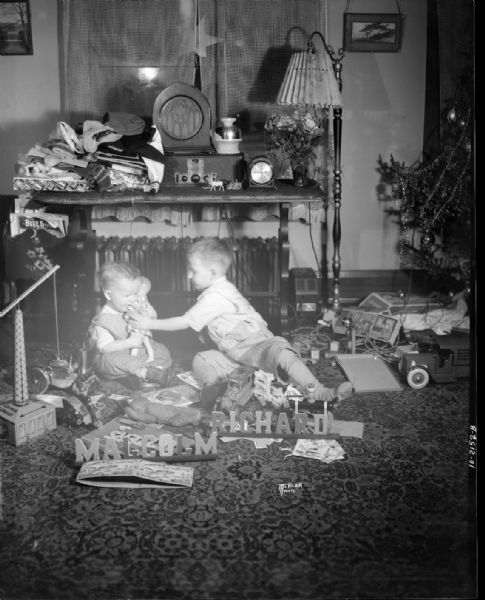 Richard and Malcom McVicar sitting on the floor playing with toys. In front of the boys are small signs that read: "Malcom" and "Richard." There is a Christmas tree on the far right.