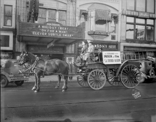 View across street towards an advertisement for the movie "Third Alarm" on the side of a fire wagon with two fire fighters being pulled by horses in front of the Strand Theatre, at 16 E. Mifflin Street.  Also shows marquee with "A Holiday treat, 'Oh For A Man,' Clever, Subtle, Smart." Shows Barton's Dresses, at 18 E. Mifflin Street, and Hughes Rose Bastic Beauty Shoppe, at 18 E. Mifflin Street.