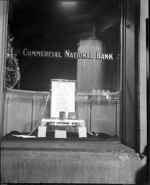 Commercial National Bank window with sign saying: "Is your money canned? Money kept at home is in danger of loss and destruction." Display shows can and toaster on top of two-burner gas stove. On the left is part of a Christmas tree.