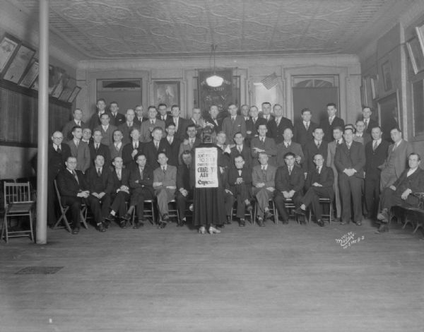 View towards a man dressed as a woman, with his back turned, standing in front of a group of American Legion men posing in a room at the G.A.R. Hall. The man is wearing a sign on his back that is advertising "Charlie's Aunt" playing at the Capitol Theatre.