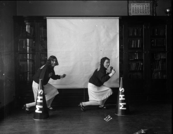 Two Middleton High School female cheerleaders in uniform. On the floor in front of them are two megaphones with the letters: "MHS." A screen has been pulled down as a backdrop over the glass doors of built-in bookcases along the back wall.