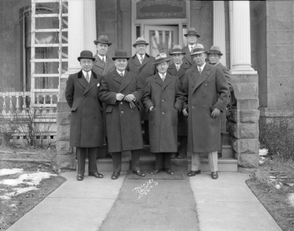 Group portrait of Governor Philip La Follette and eight industrialists standing in front of the Governor's Residence, 125 East Gilman Street. First row left to right: L.R. Smith of A.O. Smith, Gov. La Follette, C.W. Nash of Nash Motors, Co., and C.R. Messinger, of the Chain Belt Co. In the second row, left to right: Prof. John R. Commons, of the UW Economics dept., Assemblyman Harold M. Groves, Gen. Otto Falk, Allis-Chalmers Co., L.R. Clausen, of the J.I. Case co. standing in the rear, S.B. Way, of the Milwaukee Electric Railway and Light Co.