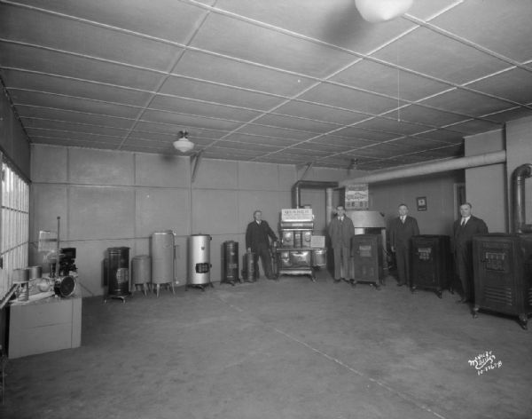 Farm Utilities Co. interior, 124 S. Butler Street. There are four men standing with water heaters, a Quaker cook stove and Quaker furnaces.