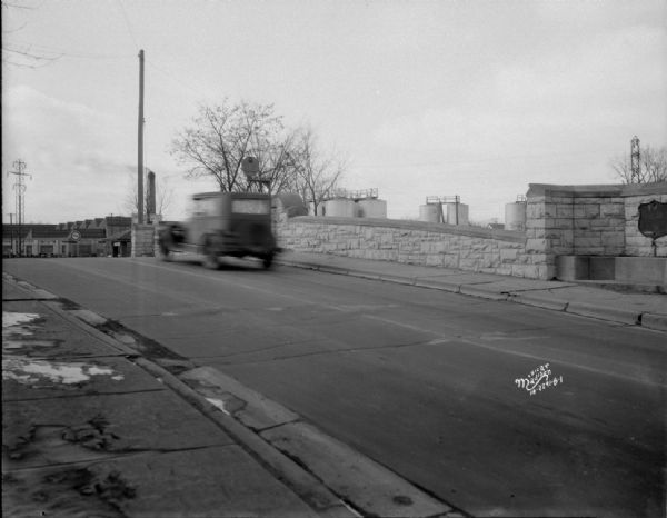 New stoplight on E. Washington Avenue bridge over the Yahara river, facing West. A truck is going over the bridge. On the other side of the bridge are industrial buildings with a sign for Fuller & Johnson Mfg. Co. Storage tanks are behind the bridge, and one of them has a sign for Deep-Rock Gasoline. There is a plaque on the bridge to the right.