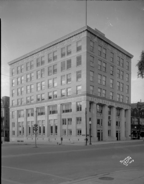 View of State Bank of Wisconsin, 1 W. Main Street. Hotel Monona is at 114 Monona Avenue, Badger Candy Kitchen is at 7 W. Main Street, E.W. Parker Jewelers is at 9 W. Main Street, and a sign painted on the side of a building reads: "Today I will eat at Cop's Cafe."