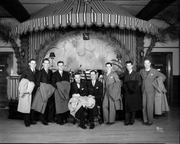 The men in the Chanticleer band are posing in front of the stage in the ballroom holding their coats.