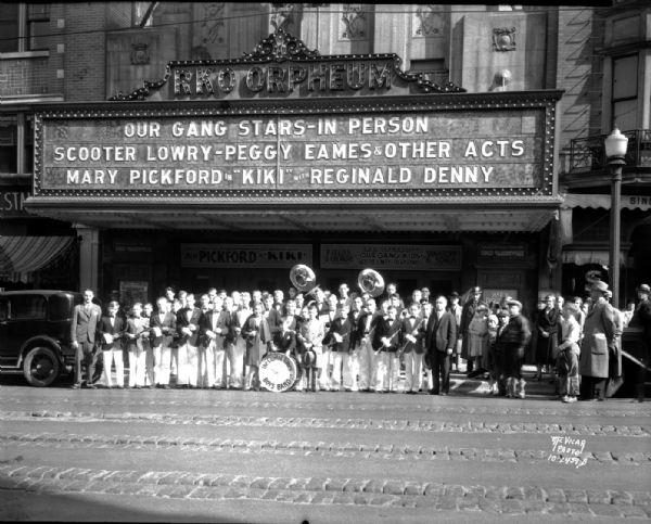 Capitol Times Boys Band in uniform with intruments standing in front of the Orpheum Theatre, 216 State Street. Marquee says "Our Gang Stars — in Person, Scooter Lowry — Peggy Eames & other acts, and Mary Pickford in 'Kiki' with Reginald Denny."
