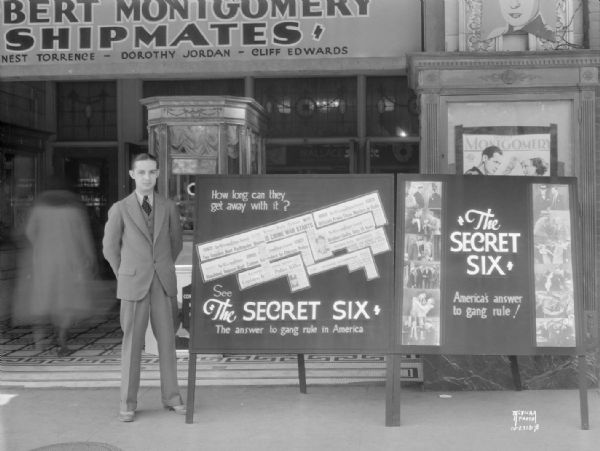 Man standing beside advertising signs for Wallace Beery in "The Secret Six — The answer to gang rule in America." Taken in front of the Strand Theatre.