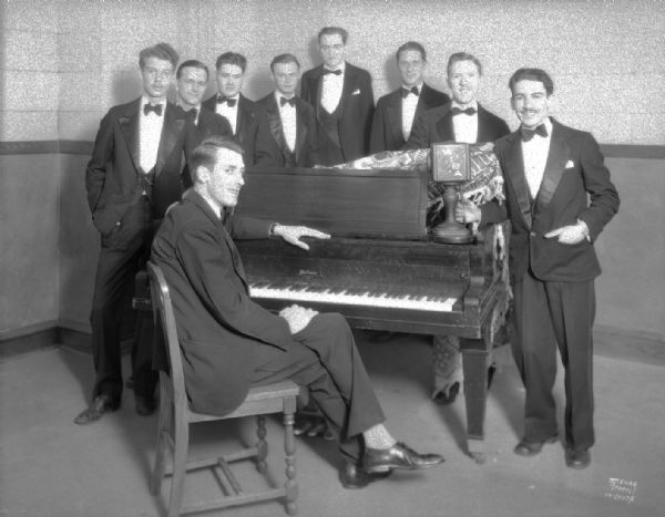 WIBA radio orchestra, Al Thompson's Esther Beach band, in tuxedos, standing around a piano with Bob Hartman sitting at the piano. Standing left to right: Norman Phelps, Earl Smith, Norman Kingsley, Arthur Kruetz, Dan McManman, James Pashak, Rollo Laylan, and Cliff Gomon.