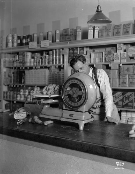 View of Anglidle scale in Dodgeville store showing grocer weighing bananas. Merchandise is on display on the counter and on shelves in the background.