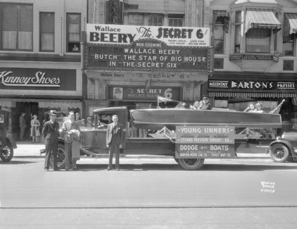 Madison Motor Car Co. — Tracy Boat Co. Dodge boat, mounted on the back of a truck, with 5 passengers and a sign advertising "Young Sinners." In front of Strand Theatre marquee featuring Wallace Beery in "The Secret 6." Also shows Kinney Shoes, 14 E. Mifflin Street, Barton's Dress Shop 18 E. Mifflin Street, Rose Bastic Beauty Shoppe, 18 E. Mifflin Street.