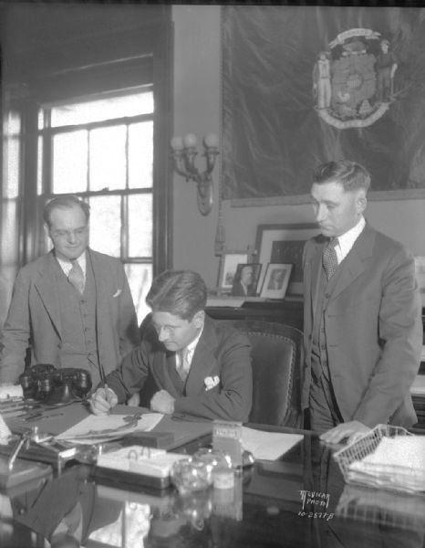 Governor Philip La Follette in the governor's office, signing extradition papers for George W.E. Perry aka "Jiggs" "Blue Beard," with Samuel Becker, executive counsel for the governor, on the left and Edmund Drager, Vilas County District Attorney, on the right.