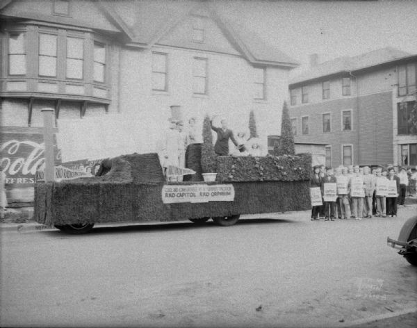 RKO parade float, with people on the float and ushers standing beside, advertising "cool and comfortable as a summer vacation" for the Orpheum and Capitol theaters. Partially visible is a Coca-Cola sign painted on the side of a building on N. Murray St. just south of University Ave.