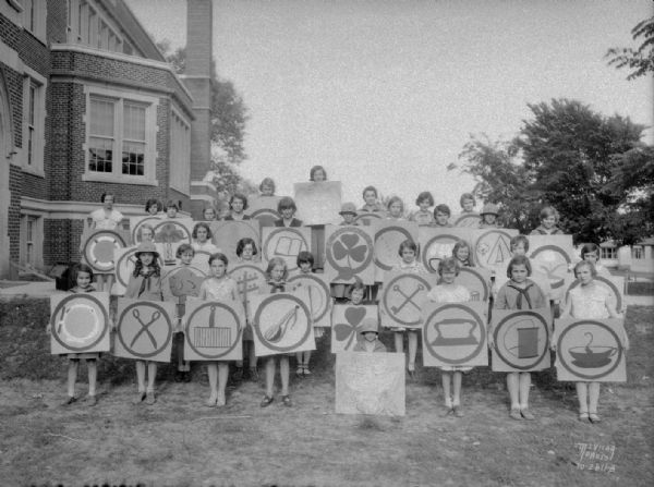 Group portrait of the Lincoln School Girl Scout Troop, holding enlarged replicas of badges earned by Dorothea Desormeaux, which earned her the Golden Eaglet award, standing outside of Franklin School, 305 W. Lakeside Street.