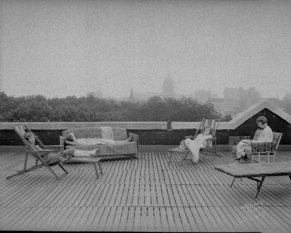 Four University of Wisconsin-Madison coeds sunbathing on the roof of Ann Emery Hall, 265 Langdon Street, an independent women's dormitory named for the first Dean of Women (1897-1900) Ann Emery Allinson (1871-1932). The Wisconsin State Capitol and the Madison skyline are in the distance.
