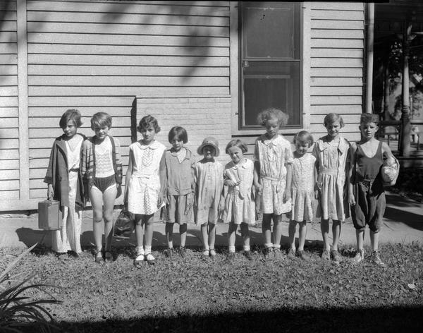 10 kids leaving for Kiddie Camp lined up in front of a building. They are wearing dresses, bathing suits, and coats.