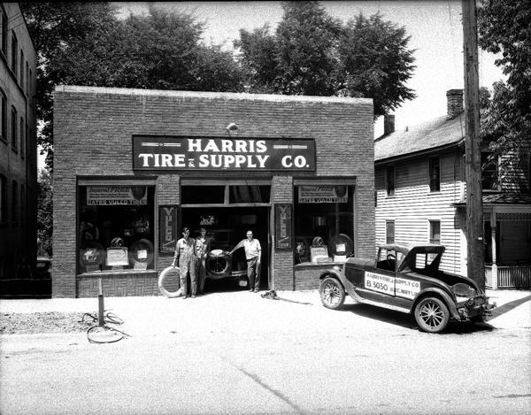 Three men standing in the doorway of Harris Tire & Supply Co. 312 E. Mifflin Street, with signs for gates, Vulco tires and roadster automobile.