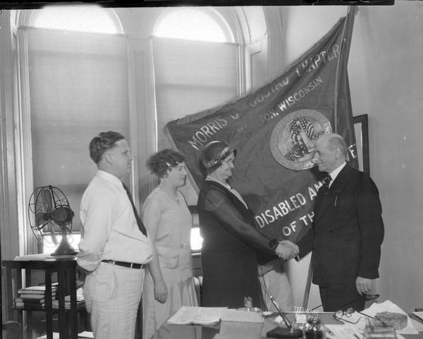 Mayor Albert Schmedeman shaking hands with Mrs. Togstad, in front of the Morris O. Togstad Chapter 2 Disabled American Veterans of the World War flag, with Roy T. and Nina Westbury looking on.